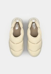 Des chaussures à enfiler de couleur crème avec un extérieur rembourré, des détails cousus et une semelle texturée. Logo de la marque visible sur la semelle intérieure et le talon.