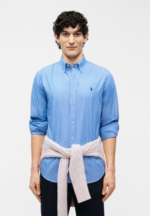 Jeune homme portant une chemise à rayures bleues à boutons avec les manches retroussées, et un pull rose clair en maille torsadée noué autour de la taille.