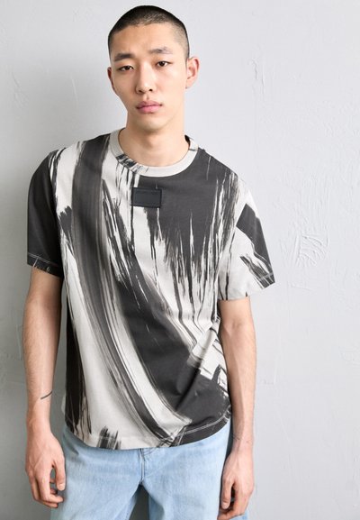 Fekete és szürke absztrakt mintájú póló, kerek nyakkivágással és a mellkason található textúrázott fekete címkével, világoskék farmernadrággal párosítva.