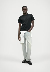 Svart t-skjorte med hvit logo, lyseblå jeans og svarte sko. En hvit hettegenser er knyttet rundt livet, med grafisk detalj.