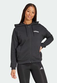 Svart zip-up hoodie med dragsko på huven, mjuk textur och vit "adidas" logotyp. Kombinerad med glansiga svarta leggings med vita ränder.