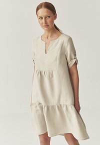 Robe en lin beige clair avec des manches courtes retroussées, un décolleté en V et un buste froncé, présentant un ourlet évasé et une texture subtile.