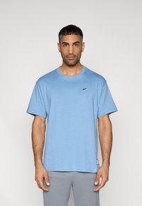 Ανοιχτό μπλε βαμβακερό t-shirt με κοντά μανίκια, στρογγυλή λαιμόκοψη και μικρό μαύρο λογότυπο στο στήθος· συνδυασμένο με γκρι παντελόνι.