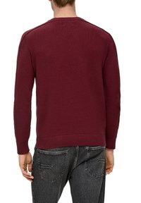 Bordeauxrotener Pullover mit strukturiertem Strick, Rundhalsausschnitt und geripptem Saum sowie Bündchen. Kombiniert mit dunklen Jeans mit Gesäßtaschen.