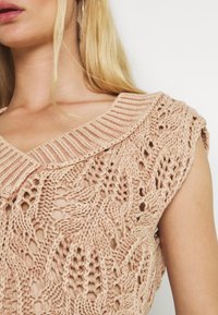 Stickad topp i ljus beige med ett strukturerat, öppet mönster. Har en ribbad, bred krage och korta ärmar. Mjuk garnmaterial.