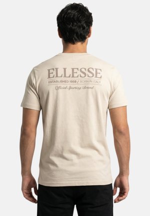 Homme aux cheveux bouclés foncés portant un t-shirt beige Ellesse avec un logo et un texte dans le dos, associé à un pantalon noir, debout, dos tourné.