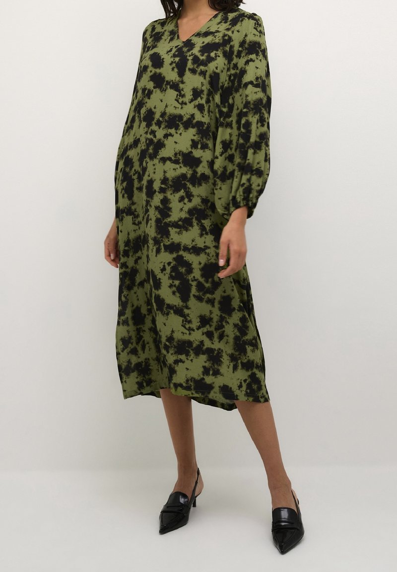 Femme portant une robe ample mi-longue vert olive avec des taches noires abstraites et des manches ballon, associée à des escarpins slingback noirs à bout pointu.