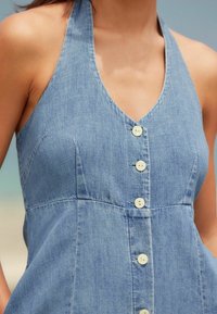 Robe en denim bleu clair avec un décolleté en V, des bretelles halter et cinq boutons blancs sur le devant. Le tissu a une finition texturée.