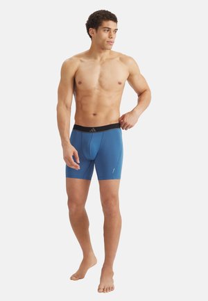 Atletische man die blootsvoets staat en blauwe boxershorts met een zwarte tailleband draagt, terwijl hij met zijn rechterhand de tailleband verstelt.