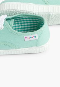 Scarpe di tela verde chiaro con suola e dettagli in gomma bianca. All'interno presentano un motivo a scacchi. L'etichetta laterale mostra "pisamonas" in multicolore.