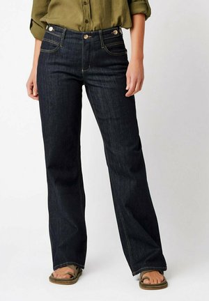 Person trägt dunkelblaue Jeans mit geradem Bein, olivgrünes Knopfhemd mit aufgekrempelten Ärmeln und grüne Sandalen.