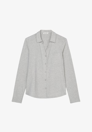 Mujer con camisa gris claro abotonada, falda de pana azul hasta la rodilla con cinturón y botas beige hasta la mitad de la pantorrilla con suela gruesa, de pie contra una pared blanca.
