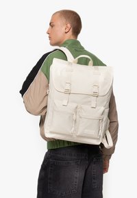 Zaino bianco in tela con flap, due tasche frontali e spallacci regolabili, caratterizzato da una texture che suggerisce durata e utilizzo all'aperto.