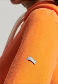 Sudadera naranja con una textura suave, un bolsillo canguro y un parche de logo azul en la manga. La capucha está forrada con un material contrastante.