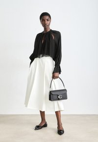 Blusa nera trasparente con scollo a cravatta, gonna midi bianca plissettata, borsa nera con accessori in argento e ballerine nere a punta.
