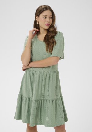 Kobieta o długich brązowych włosach nosi jasną zieloną sukienkę z ażurowym wzorem, złote kolczyki koła i bransoletki, pozuje z założonymi rękami, patrząc na bok.