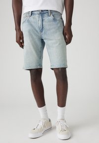 Muž oblečený v světle modrých denimových šortkách, bílých ponožkách po holeně a béžových teniskách stojí proti jednoduchému světlému pozadí.