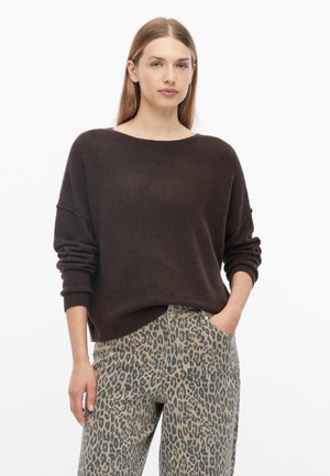 Jonge vrouw met steil haar, gekleed in een donkerbruine trui en een broek met luipaardprint, staand met één hand in de zak tegen een witte achtergrond.