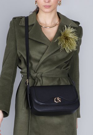 Borsa a tracolla nera in pelle con hardware dorato, forma rettangolare, superficie texturizzata e chiusura a pattina curvata, indossata sopra un cappotto verde oliva.
