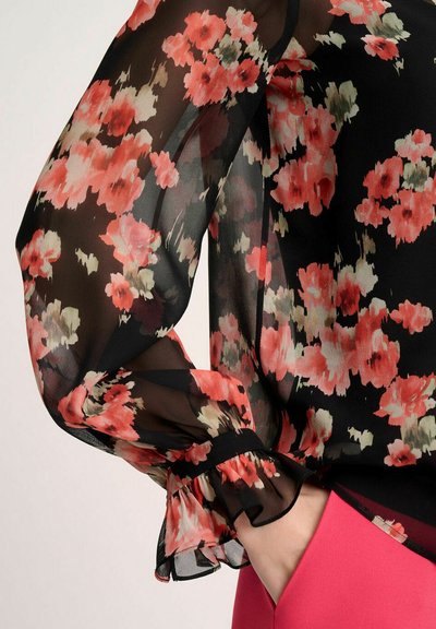Blusa negra transparente con patrón floral en rosa y verde, con puños acampanados y corte holgado. La tela es ligera y transparente.