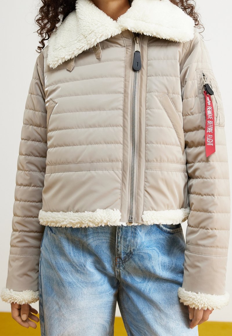 Beżowa kurtka puchowa z białym sztucznym futrzanym kołnierzem i mankietami, z przodu z zamkiem błyskawicznym oraz boczną kieszenią z czerwoną metką.