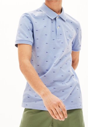 Mies, jolla on vaaleansininen Lacoste-pikeepaita, jossa pieniä tummansinisiä krokotiililogoja, ja oliivinvihreät housut, seisomassa valkoista taustaa vasten.