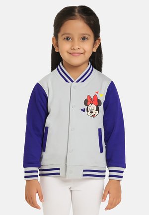 Una giovane ragazza sorride, indossando una giacca varsity grigio chiaro e viola con Minnie Mouse e cuori sul petto, abbinata a pantaloni bianchi.