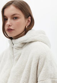 Felpa con cappuccio in pelliccia sintetica color crema, con colletto con zip e vestibilità rilassata. Presenta una consistenza morbida e un cappuccio per garantire calore. Sottogiacca marrone con collo alto.