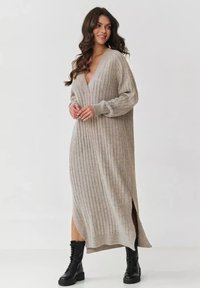 Długa, prążkowana sukienka dzianinowa w jasnym beżu z głębokim dekoltem w kształcie V i bocznymi rozcięciami. Połączona z czarnymi sznurowanymi botkami bojowymi.