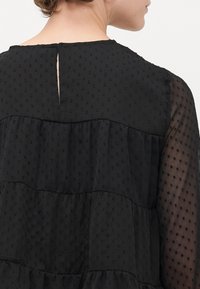 Frau trägt eine schwarze strukturierte Bluse mit transparenten, gepunkteten langen Ärmeln und einem kleinen Schlüsselloch-Ausschnitt am Rückenausschnitt.
