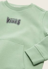 Sudadera verde menta con cuello redondo, puños acanalados y un bolsillo frontal. Presenta un bordado negro que dice "AWESOME VIBES". Textura suave.