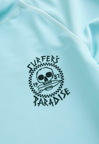 Lichtblauwe rash guard gemaakt van zacht materiaal met een zwart grafisch ontwerp van een schedel en een surfplank, tekst "SURFER'S PARADISE" en "1982."