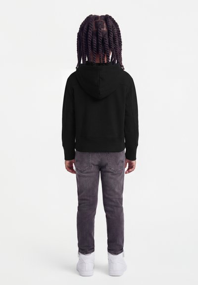 Παιδί με πλεξούδες, που φοράει μαύρο φούτερ με κουκούλα, ξεθωριασμένα γκρι τζιν και λευκά αθλητικά παπούτσια, στέκεται γυρισμένο με την πλάτη σε απλό φόντο.