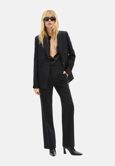 Black tailored suit with a double-breasted blazer and wide-leg trousers. Satin shirt underneath, paired with black pointed heels and sunglasses.