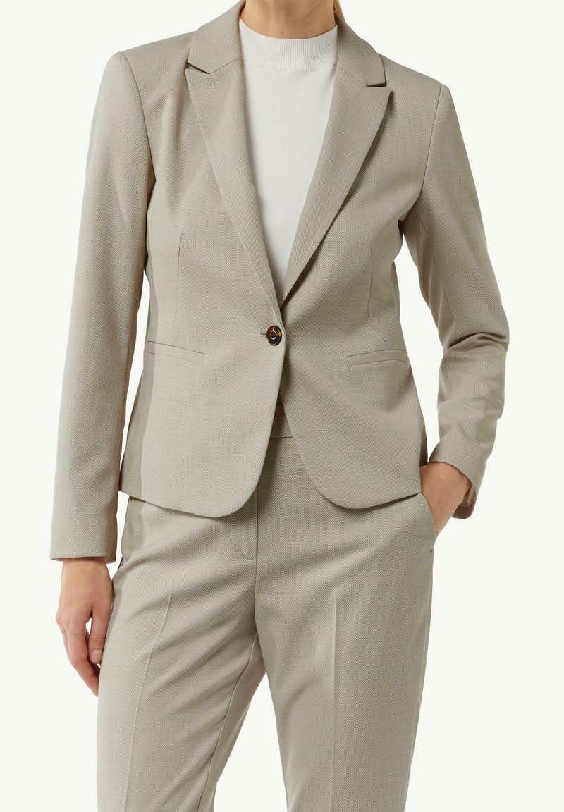 Femme portant un tailleur beige sur mesure avec un blazer à un seul bouton et un pantalon assorti, associé à un top blanc à col montant.