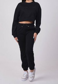 Kobieta ubrana w czarny krótki sweter i dresy, w białych i beżowych butach sportowych, stojąca na tle jednolitego jasnego tła.