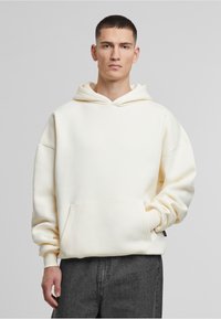 Cremefarbene Kapuzenjacke aus weichem Material mit einer Fronttasche, lässiger Schnitt und gerippten Bündchen. Getragen mit dunkelgrauen Hosen.