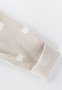 Tessuto in cotone beige con una superficie testurizzata e motivi a forma di corona bianchi. Presenta un ampio polsino a coste a un'estremità.