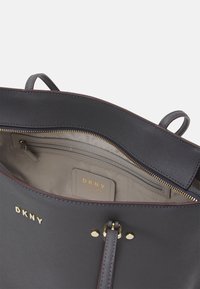 Torba tote DKNY o gładkim szarym wykończeniu, złotym suwaku i wewnętrznych przegródkach. Ma podszewkę z tkaniny i metkę z wytłoczonym logo.