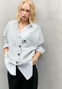 Svetlošedý pletený cardigan s rebrovaným golierom, piatimi tmavohnedými gombíkmi, kombinovaný s bielou prúžkovanou košeľou. Hladká textúra, uvoľnený strih.