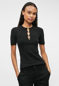 Svart ribbestrikket topp med kort ermer og rund hals, med fire gullknapper med preget tekst på midten foran.