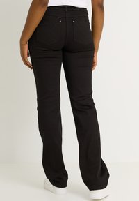 Persona vestida con jeans de corte acampanado negros y zapatillas blancas, de pie contra un fondo claro y liso, vista desde atrás.