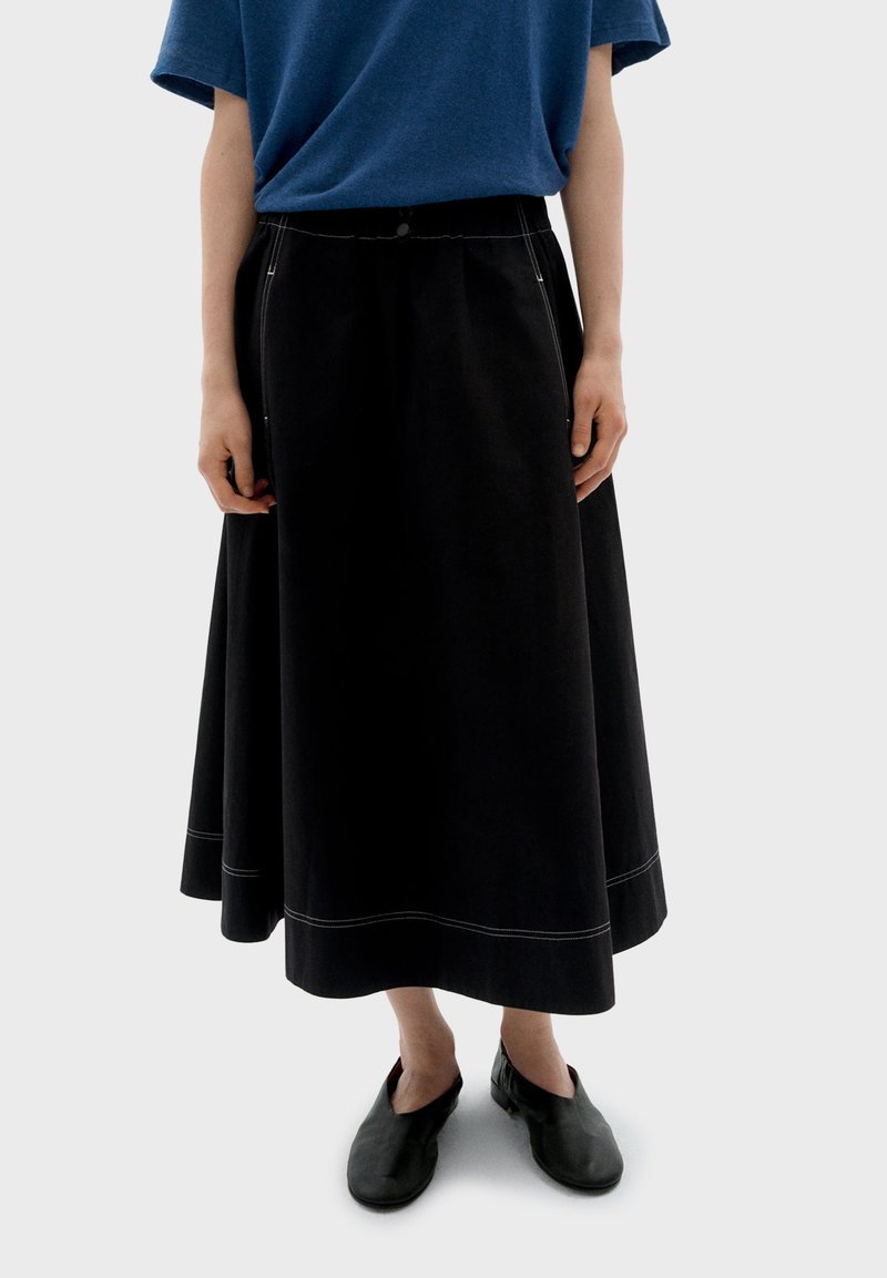 Jupe midi noire à coupe évasée, avec des coutures contrastantes et une fermeture à boutons à la taille, associée à des chaussures noires à enfiler.