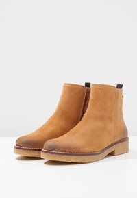 Stivaletti alla caviglia in suede marrone con zip laterale, punta arrotondata e suola marrone con texture a contrasto. Parte superiore liscia e dettagli di cucitura rinforzata.