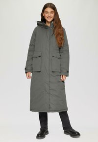 Long manteau gris rembourré avec capuche, deux poches avant et bouton au col. Un pantalon noir et des bottes noires imposantes complètent la tenue.