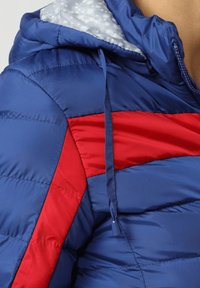 Chaqueta acolchada azul con un diseño angular rojo, capucha con cordón y un forro interior gris claro con patrón. Textura suave y brillante.