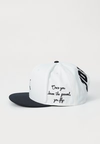 Casquette blanche avec une visière noire, présentant un texte brodé sur le côté : "Une fois que vous quittez le sol, vous volez." Le matériau est un mélange de coton.