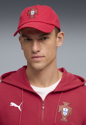 Joven con gorra de béisbol roja de Portugal y sudadera con cremallera a juego con los escudos de Puma y de fútbol de Portugal.