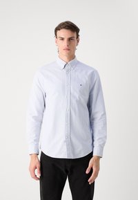 Camisa abotonada en azul claro con rayas blancas, de mangas largas y con un dobladillo redondeado. Presenta un pequeño logotipo en el lado izquierdo del pecho.