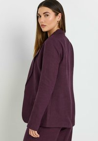 Mörk lila linnemäntel med en skräddarsydd passform, revär med snibb och sidfickor, visad i en sidovy som framhäver dess textur.